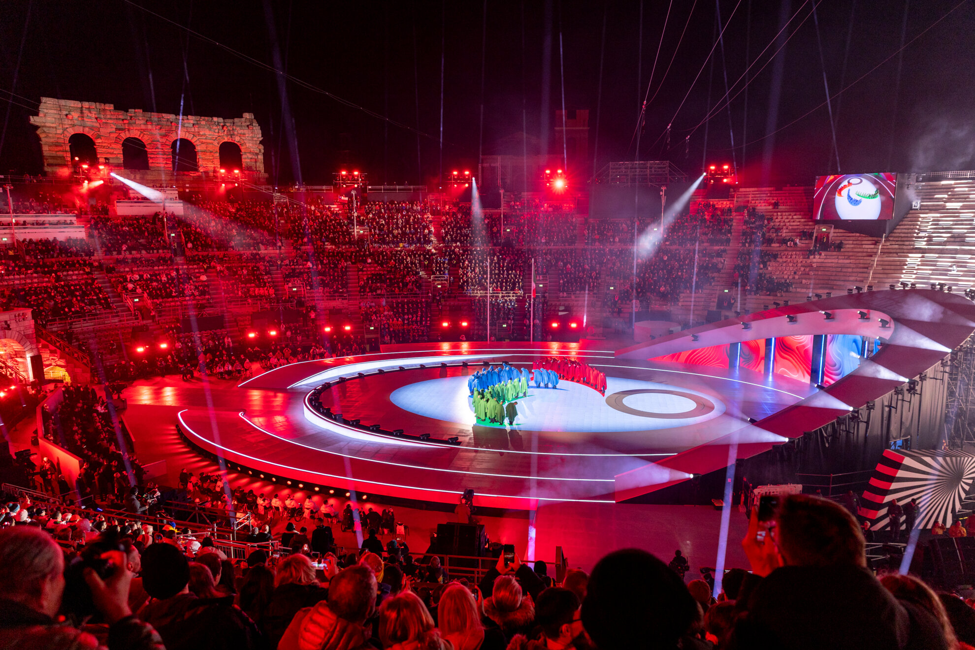 Areena hehkuu punaisena, ja keskellä lavalla sini-, puna- ja vihreäasuiset tanssijat muodostavat Agitos-merkin.